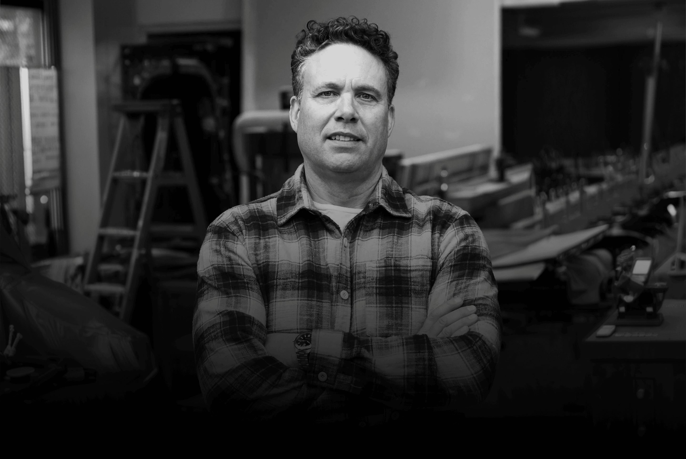 Man with arms crossed wearing a plaid shirt stands in a workshop with tools visible in the background.