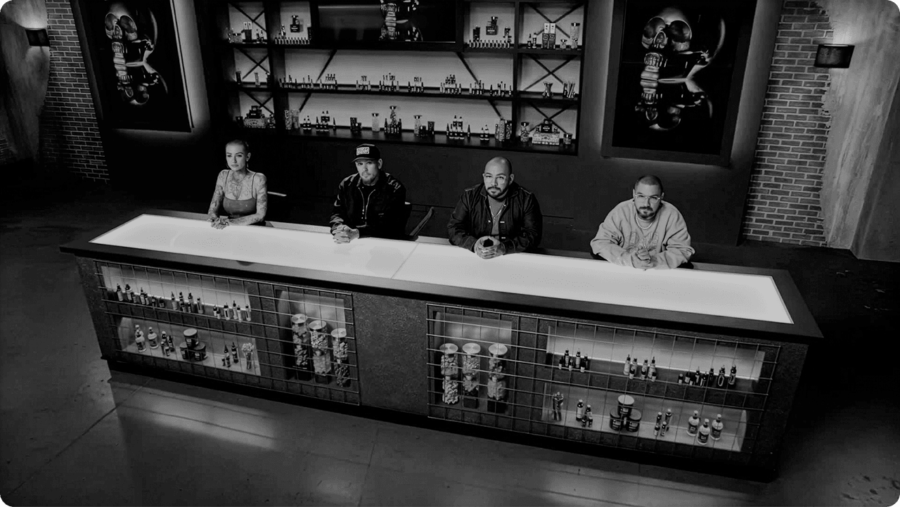 Ink Master judges seated at illuminated desk with bar backdrop