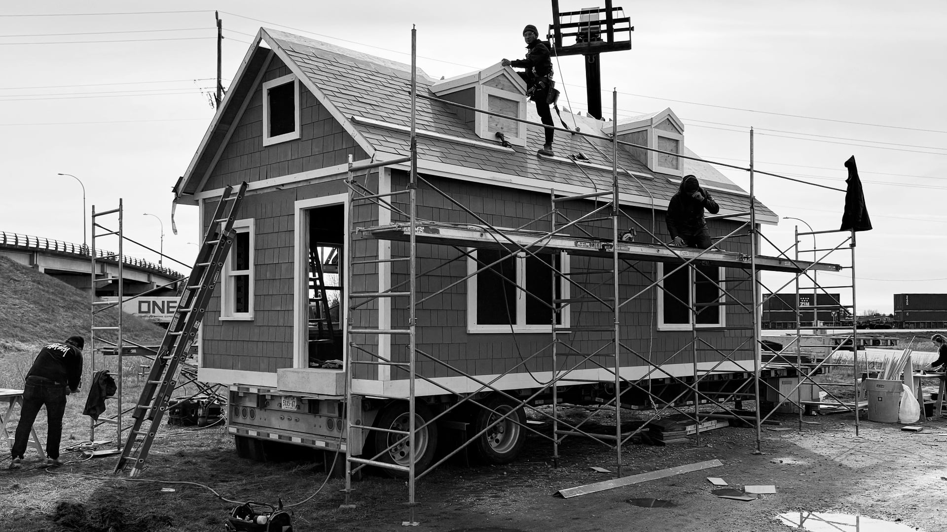 Crew on scaffolding finishing exterior of portable grey shingle cottage on flatbed trailer for Nissan commercial