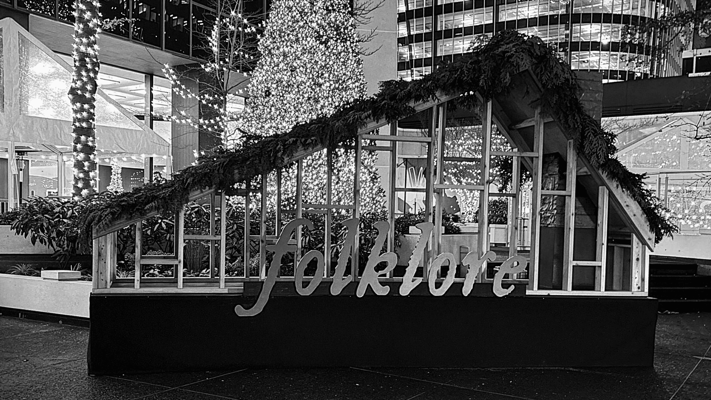 Taylor Swift Folklore cabin installation lit at night with evergreen garland and illuminated sign in urban plaza